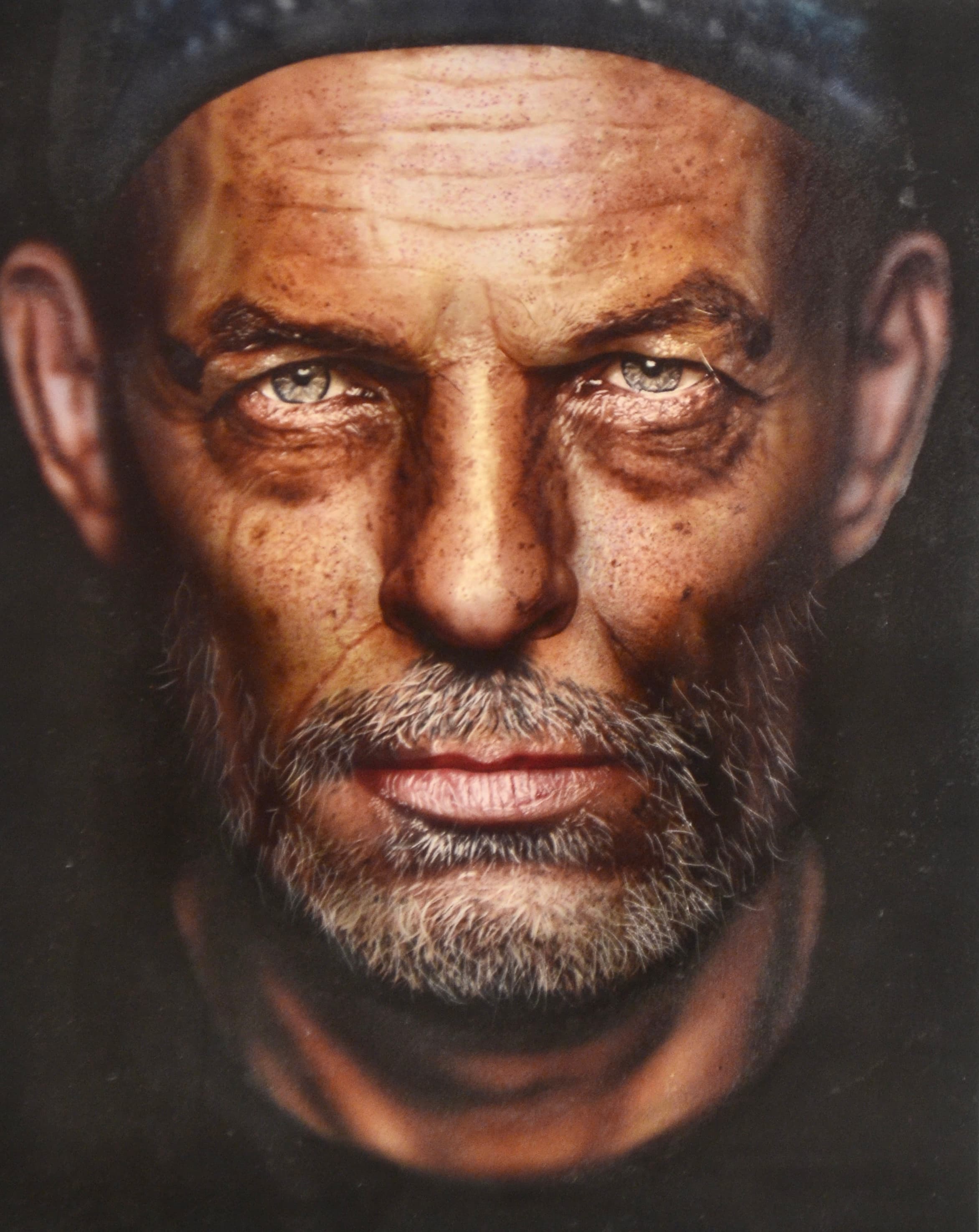 Detailed portrait of a weathered man with intense light eyes, grey beard, and dark cap.